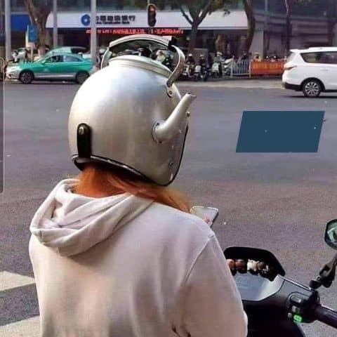 Person on a scooter wearing a metallic helmet shaped like a kettle with a spout and handle, facing a street intersection.