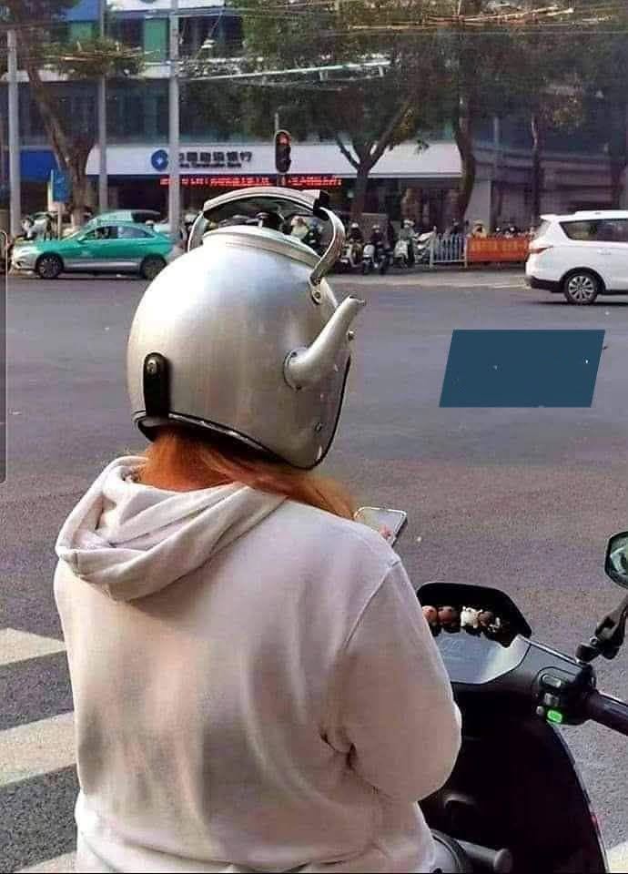 Person on a scooter wearing a metallic helmet shaped like a kettle with a spout and handle, facing a street intersection.