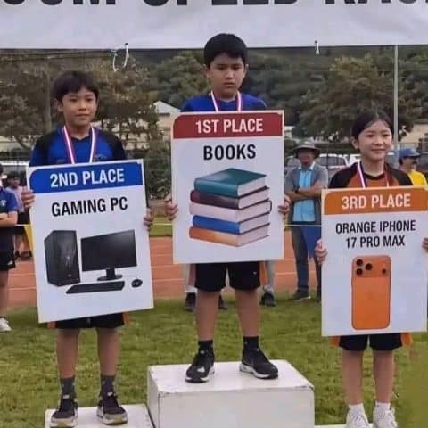 Three kids on a winners podium hold large signs showing their prizes: first place with books, second with a gaming PC, and third with an orange iPhone 17 Pro Max.