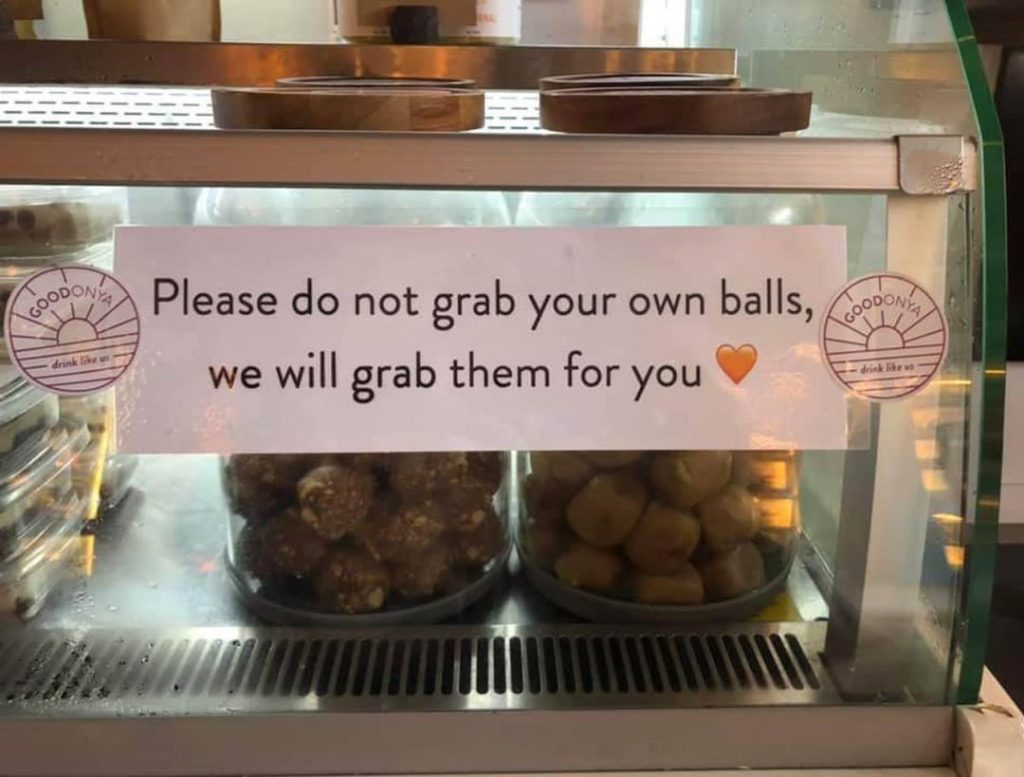 A sign on a food display case reads 'Please do not grab your own balls, we will grab them for you' with two trays of round snack balls visible inside.