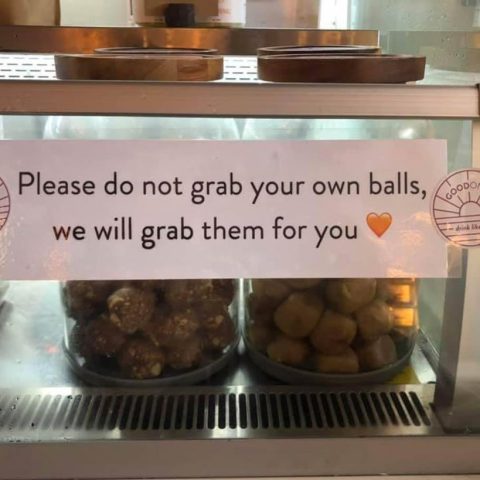A sign on a food display case reads 'Please do not grab your own balls, we will grab them for you' with two trays of round snack balls visible inside.