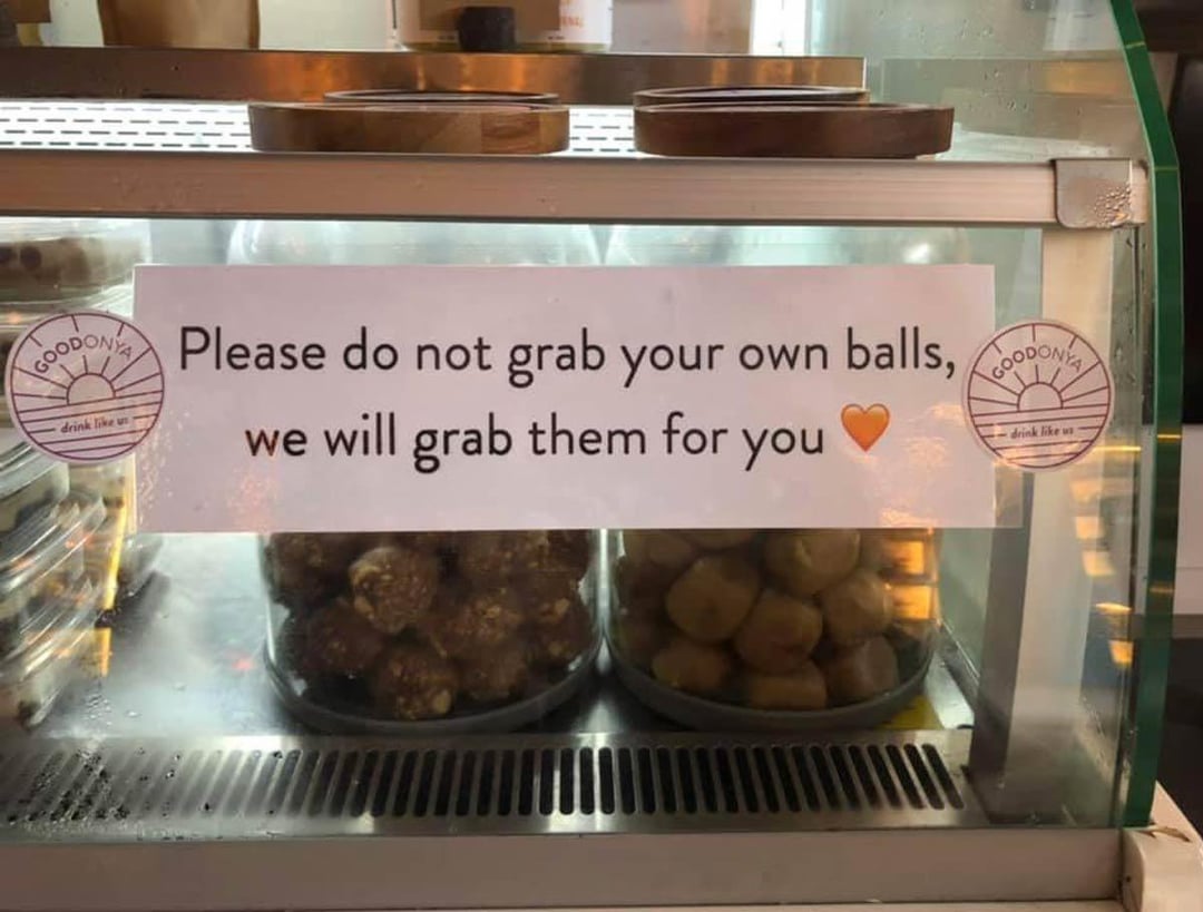 A sign on a food display case reads 'Please do not grab your own balls, we will grab them for you' with two trays of round snack balls visible inside.