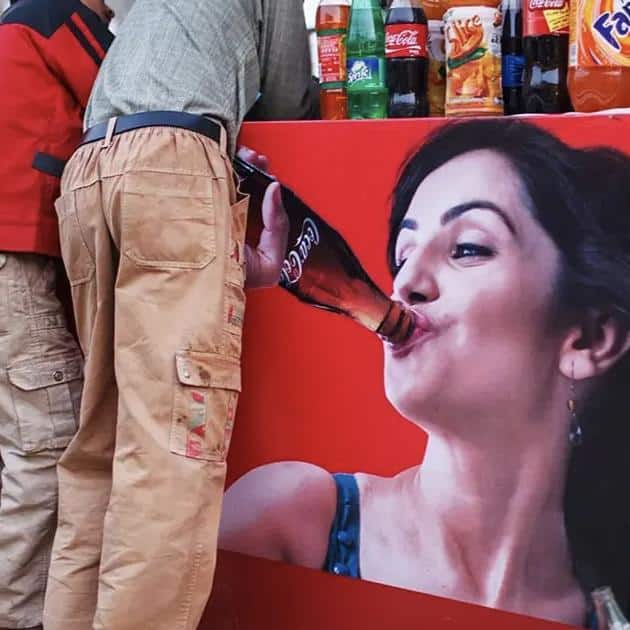 A man leaning into a counter with a large poster of a woman drinking a Coca-Cola bottle, creating the illusion that he is drinking from the bottle.