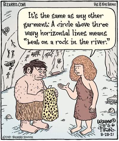 Two cavemen in a cave with drawings on the walls; one explains to the other a laundry symbol meaning 'beat on a rock in the river' while holding a spotted garment.