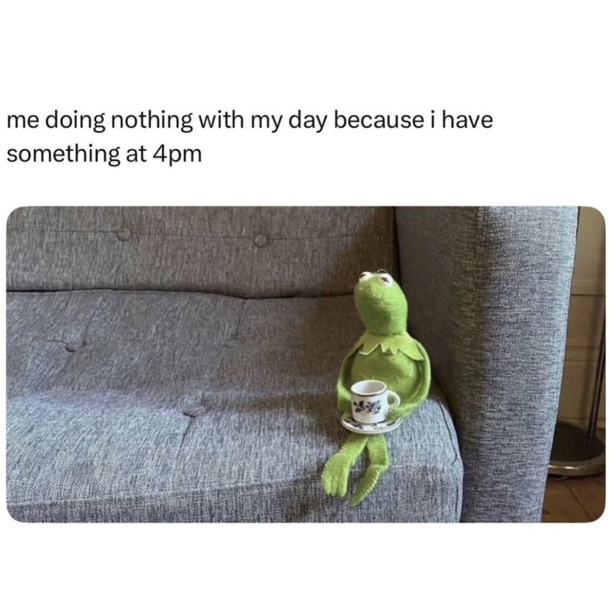 A Kermit the Frog plush toy sits alone on a grey fabric couch holding a small teacup and saucer, looking upward thoughtfully.