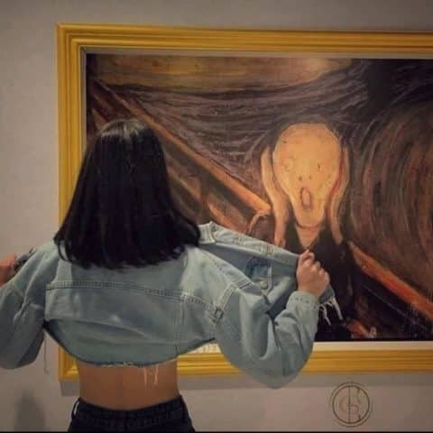 A woman with shoulder-length dark hair, wearing a cropped denim jacket, stands in front of The Scream painting, pulling her jacket open to show her back.
