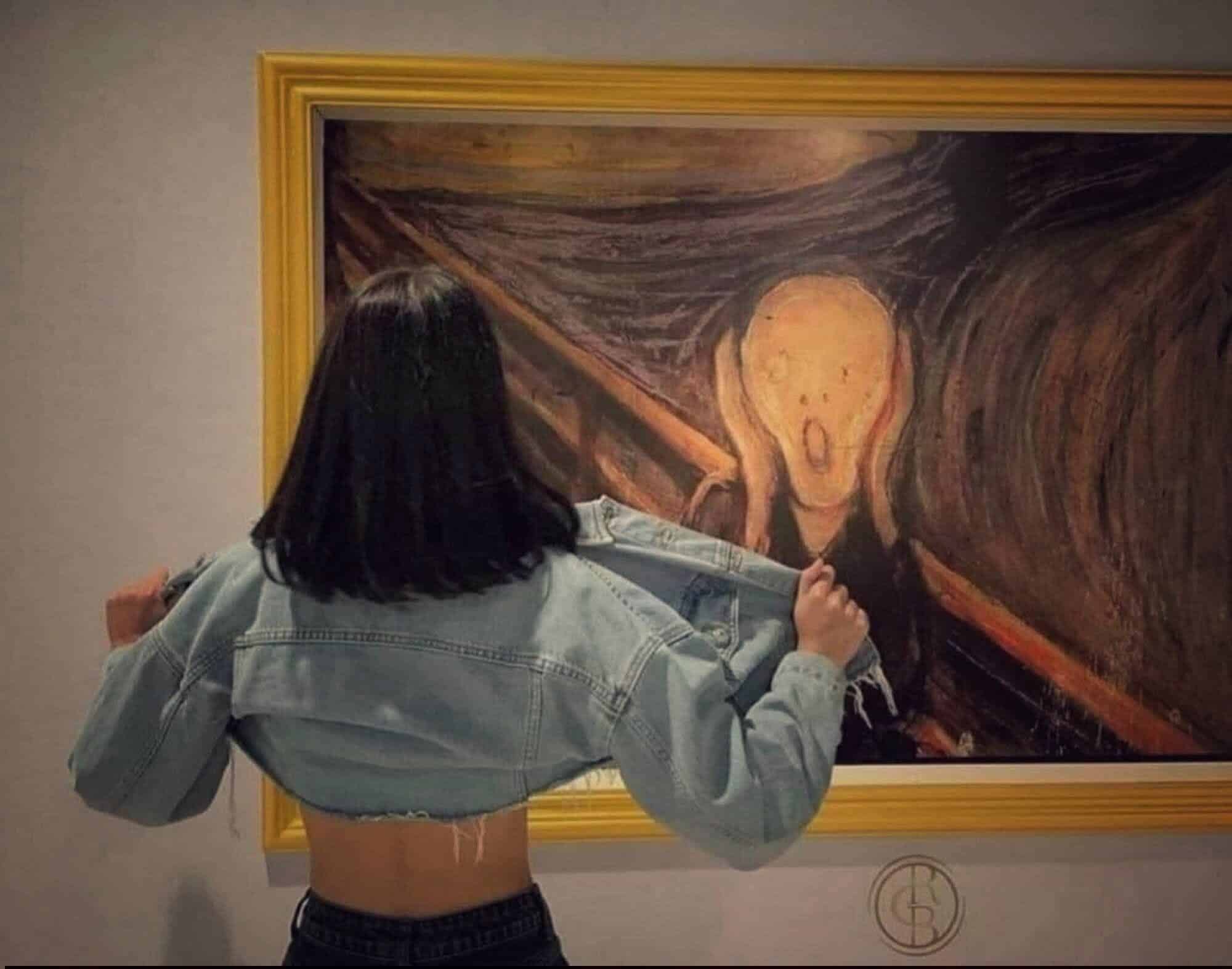 A woman with shoulder-length dark hair, wearing a cropped denim jacket, stands in front of The Scream painting, pulling her jacket open to show her back.