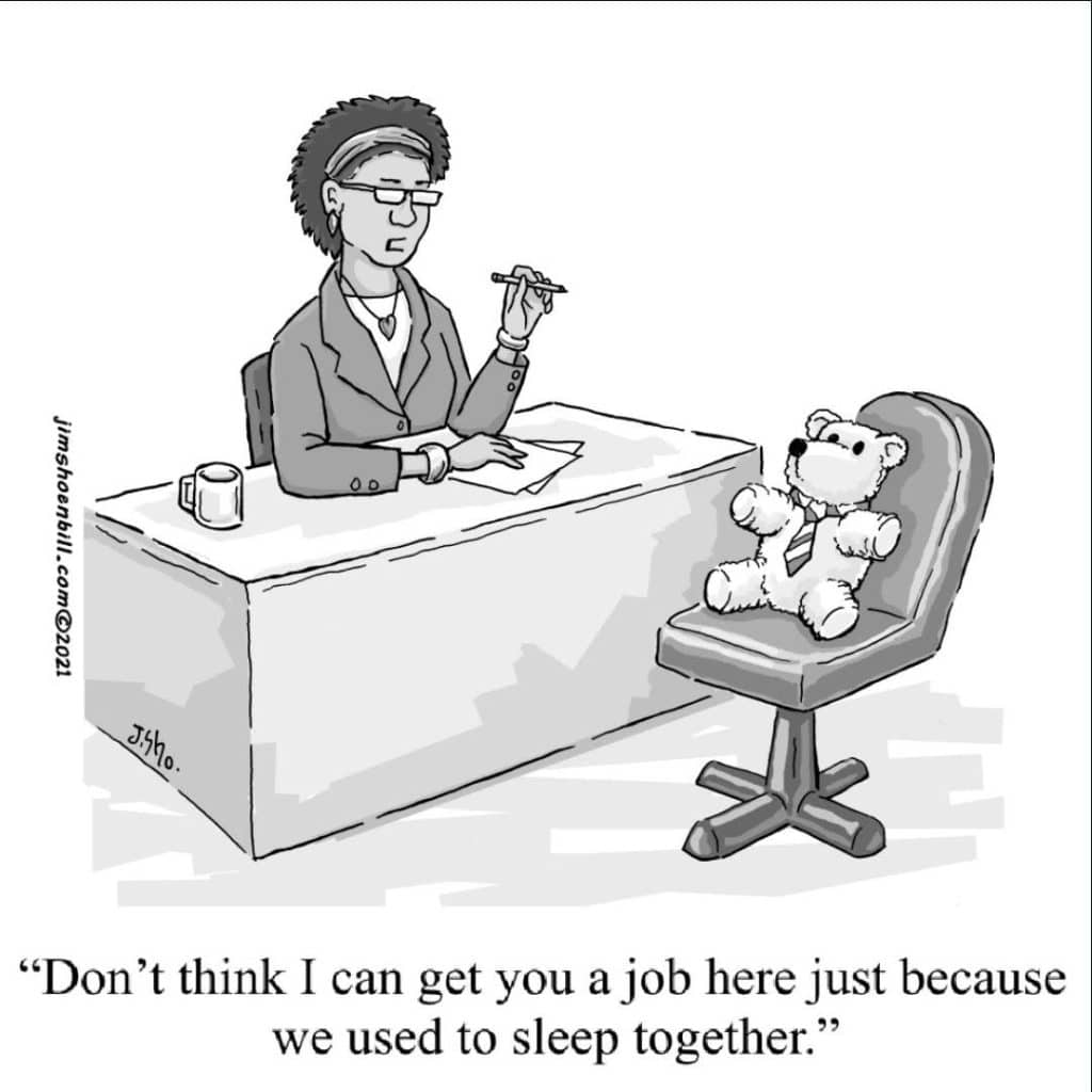 A woman sits at an office desk holding a pen, talking to a teddy bear wearing a tie seated in a chair opposite her.