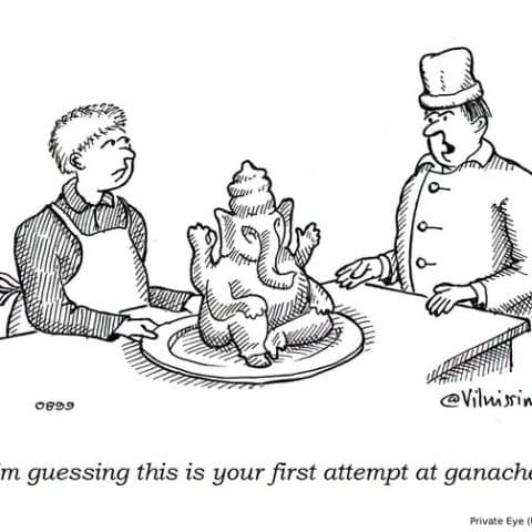 Two men stand by a table. One wears an apron and the other a chef's hat. On the table is a chocolate ganache shaped like the Hindu god Ganesha. The man in the chef's hat looks surprised and comments.