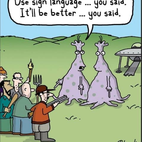 Two purple aliens with antennae stand on a grassy field. One alien has a speech bubble saying, 'Use sign language ... you said. It'll be better ... you said.' They face a mob of angry people holding weapons, with a UFO in the background.