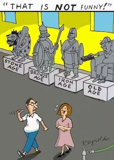 A museum exhibit has statues labeled Stone Age, Bronze Age, Iron Age, and Old Age. The first three are historic warrior figures, while the last is an elderly man sitting with a remote control, causing a man nearby to angrily say "That is not funny!"